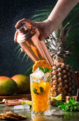 Wall Mural - Summer cocktail with vodka, pineapple juice, mango, ice. Long drink or cold mocktail. Bartender shakes copper shaker with splashes, frozen motion and flying drops. Tropical background with palm leaves