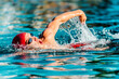 Male swimmer swimming crawl freestyle in blue water outdoor pool. Portrait of an athletic young male triathlete swimming crawl wearing a red cap and swim goggles. Triathlete training for triathlon