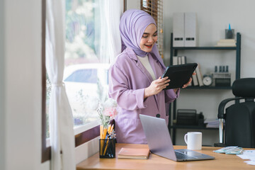 Wall Mural - A Muslim millennial businesswoman, wearing a purple hijab, working with tablet and financial report paperwork in home office. The concept of financial advising, teamwork, and accounting is depicted.