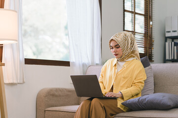 Wall Mural - A stunning Muslim millennial businesswoman wearing a yellow hijab working from her home office while sitting on a couch in her living room.