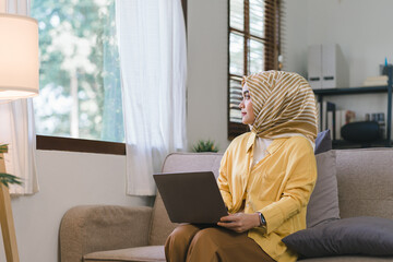 Wall Mural - A stunning Muslim millennial businesswoman wearing a yellow hijab working from her home office while sitting on a couch in her living room. Looking outside window