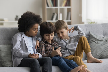 Multiethnic team of focused little kids using mobile phone together, resting on cozy couch at home. Preschool boy playing online video game on smartphone, showing screen to friends