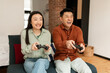 © Prostock-studio - Cool happy asian spouses sitting on couch in living room, holding joysticks and laughing, playing video games at home