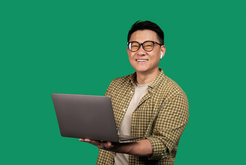 Positive asian man holding laptop computer and smiling at camera, wearing earpods and eyeglasses, green background