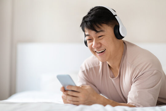 Wall Mural - Positive asian man using smartphone and headphones in bed
