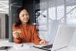 © Liubomir - Young successful businesswoman smiling and looking at laptop, Asian woman holding bank credit card and shopping online in online store, woman with laptop at workplace.