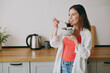 © ViDi Studio - Young fun minded dreamful happy housewife woman wear casual clothes look aside eat breakfast muesli cereals with milk fruit in bowl cooking food in light kitchen at home alone. Healthy diet concept.