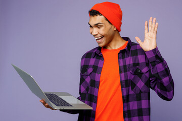 Side view young IT man of African American ethnicity in casual shirt orange hat hold use work on laptop pc computer waving hand isolated on plain pastel light purple color background studio portrait