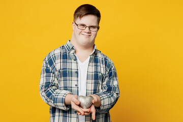 Young smiling happy man with down syndrome wear glasses casual clothes look camera hold in palms silver heart toy isolated on pastel plain yellow color background. Genetic disease world day concept.