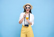 Happy smiling young asian woman tourist in summer hat standing with camera and drinking orange juice isolated on blue studio background