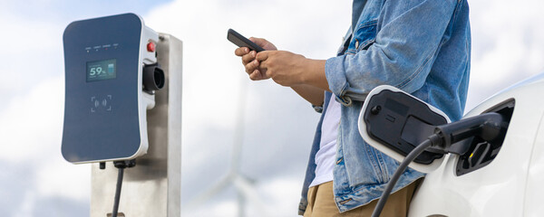 Progressive man with his electric car, EV car recharging energy from charging station on green field with wind turbine as concept of future sustainable energy. Electric vehicle with energy generator.