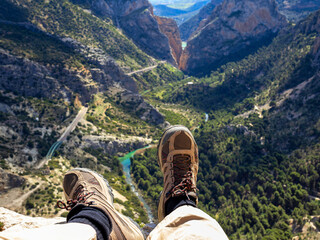 Two feet in hiking boots dangling in a ravine. In the background a valley with mountains and river.