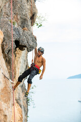 Young Asian man climber climbing on rocky coastline at tropical island in sunny day. Handsome guy enjoy outdoor active lifestyle and extreme sport training mountain climbing on summer holiday vacation