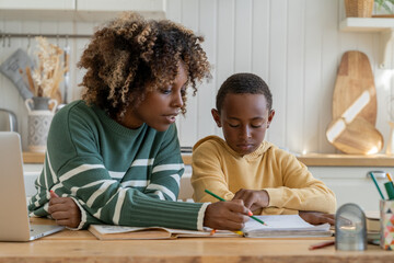 Focused biracial mother explaining difficult school task to attentive son for effective learning. African American personal tutor woman and little schoolboy sit at desk in home kitchen study together