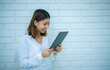 © Digital Art Studio - Pretty asian business woman stands sideways and looks at the tablet she is holding with a smile with white bricks in the background, Digital marketing.