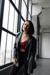 © Tetiana Romaniuk - A girl with black hair, red lipstick and a leather jacket stands near a window