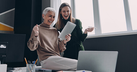 Wall Mural - Two excited women looking at digital tablet and gesturing while working in office together