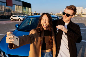 A man and a woman stand in front of their new car outside. A couple takes a selfie on a smartphone