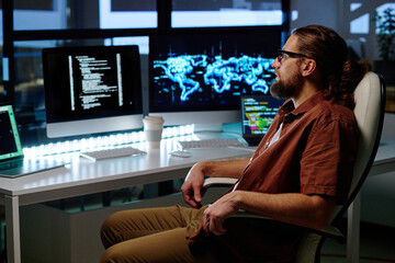 Wall Mural - Young serious IT programmer sitting in armchair by workplace in front of several computers with coded data on screens late in the evening