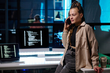 Young IT engineer consulting client on mobile phone while standing by workplace with desktop computers with coded data on screens