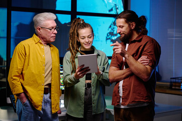 Wall Mural - Group of three programmers discussing points of presentation at working meeting in dark office while standing against workplace with computers