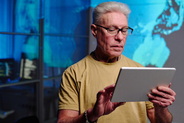 Wall Mural - Serious mature businessman with grey hair using digital tablet while standing in frnt of camera in dark office and preparing presentation