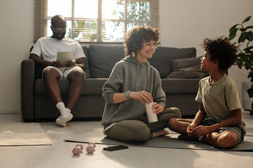 Poster - Happy young woman in activewear and her adorable son sitting on mat in living room and chatting while enjoying break after morning workout