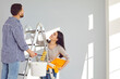 © Studio Romantic - Family couple doing renovations, painting walls and whitewashing ceiling at home. Happy young man and woman standing on step ladder, holding paint can, tray, and rollers, looking up, and smiling