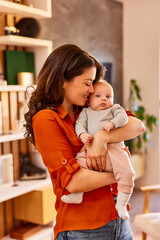 A pretty woman carrying her cute baby girl in her arms and standing at home.