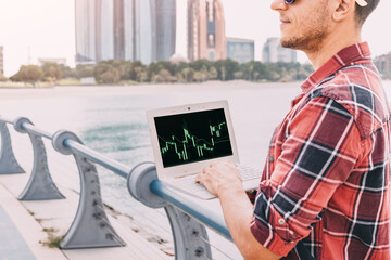 Young man working on laptop with stock market exchange and cryptocurrency online graph and chart, against skyline of Abu Dhabi downtown city
