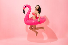Beautiful Smiling Asian Woman In Summer Bikini Swimsuit Jumping With Flamingo Swim Ring In Isolated Pink Color Studio Background