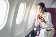 Young Asian attractive business woman passenger sitting on business class luxury plane while wearing wireless headphone and using smart phone mobile for relax during the flight