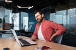 © Liubomir - Back pain at the workplace. A young male programmer, freelancer sits in the office at the table and holds his back twisted, stretched, tension pain. Tired from an overtime day.