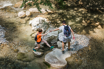 Active young hikers, man and woman walking over a mountain stream flowing among large beautiful rocks. Concept of nature exploring.