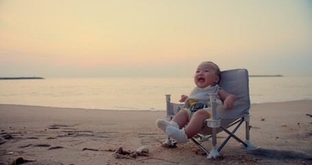 Wall Mural - happy cute adorable Asian baby infant sitting relaxing on little chair and smiling with waves on background at seaside tropical sandy beach in sunset sunrise during holiday vacation summertime