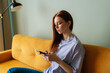 Medium shot of frustrated redhead young woman looking at smartphone screen sitting on yellow couch, received message with bad news, web surfing problem solution in internet, chatting online.