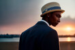 Street fashion portrait of a stylish young elegant luxury African man in a straw beige hat and coat or jacket in retro style by the lake at sunset