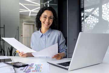 Wall Mural - Portrait of happy and successful financial woman accountant, businesswoman behind paper work with contracts and bills reports smiling and looking at camera, Hispanic woman at workplace with laptop.