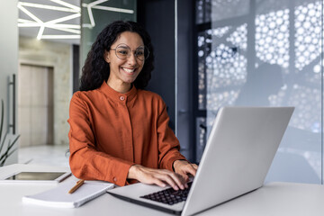 Wall Mural - Beautiful and successful hispanic business woman inside office working with laptop, office worker with curly hair smiling woman boss happy with achievement results at workplace.
