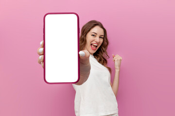 young cute woman in white t-shirt shows blank smartphone screen and rejoices in victory on pink isolated background