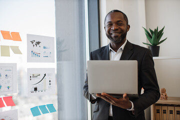 Smiling african american businessman with portable computer in hands expecting colleagues in meeting room. Financial analyst informing investment strategy using data visualizations on glass board.