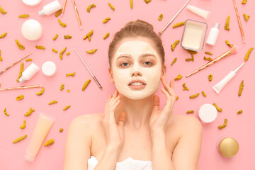 Young woman with applied turmeric mask, roots and cosmetic products on pink background