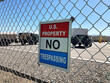 U.S. Property No Trespassing sign attached to a chain link. Blurred military equipment, vehicles in background