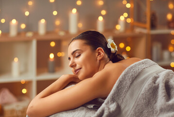 Wall Mural - Young lady getting beauty treatments at a modern spa salon. Happy, relaxed woman with closed eyes lying on the bed, with beautiful candle lights gleaming in the background. Spa, female beauty concept