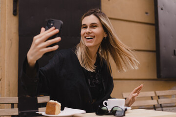 Happy woman using smartphone at cafe. Smiling blonde woman using smart phone make selfie, spends her leisure time in cafe. Blonde girl wear black shirt and make gesture it's me.