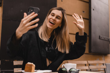 Wall Mural - Happy woman using smartphone at cafe. Smiling blonde woman using smart phone make selfie and make hello gesture waving hand, spends her leisure time in cafe, video call. Blonde girl wear black shirt.