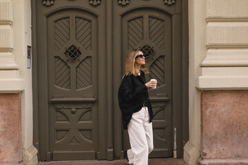 Wall Mural - Happy beautiful stylish woman having good fashion white jeans pants, black shirt walking by street, make step and holding coffee or tea in cup takeaway, good mood. Girl look happy, go away from house.