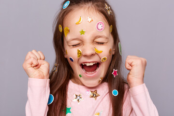 Indoor shot of overjoyed smiling cute little brown haired little girl covered with stickers posing isolated over gray background, clenched fists, celebrating her success.