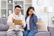 Upset Asian family, man and woman sitting together at home in living room on sofa, couple received letter mail envelope notification with bad news, unhappy with received message.