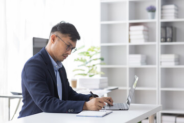 Wall Mural - Focused young adult businessman, asian man sitting at his desk making sure his company is earning enough money financial planning concept